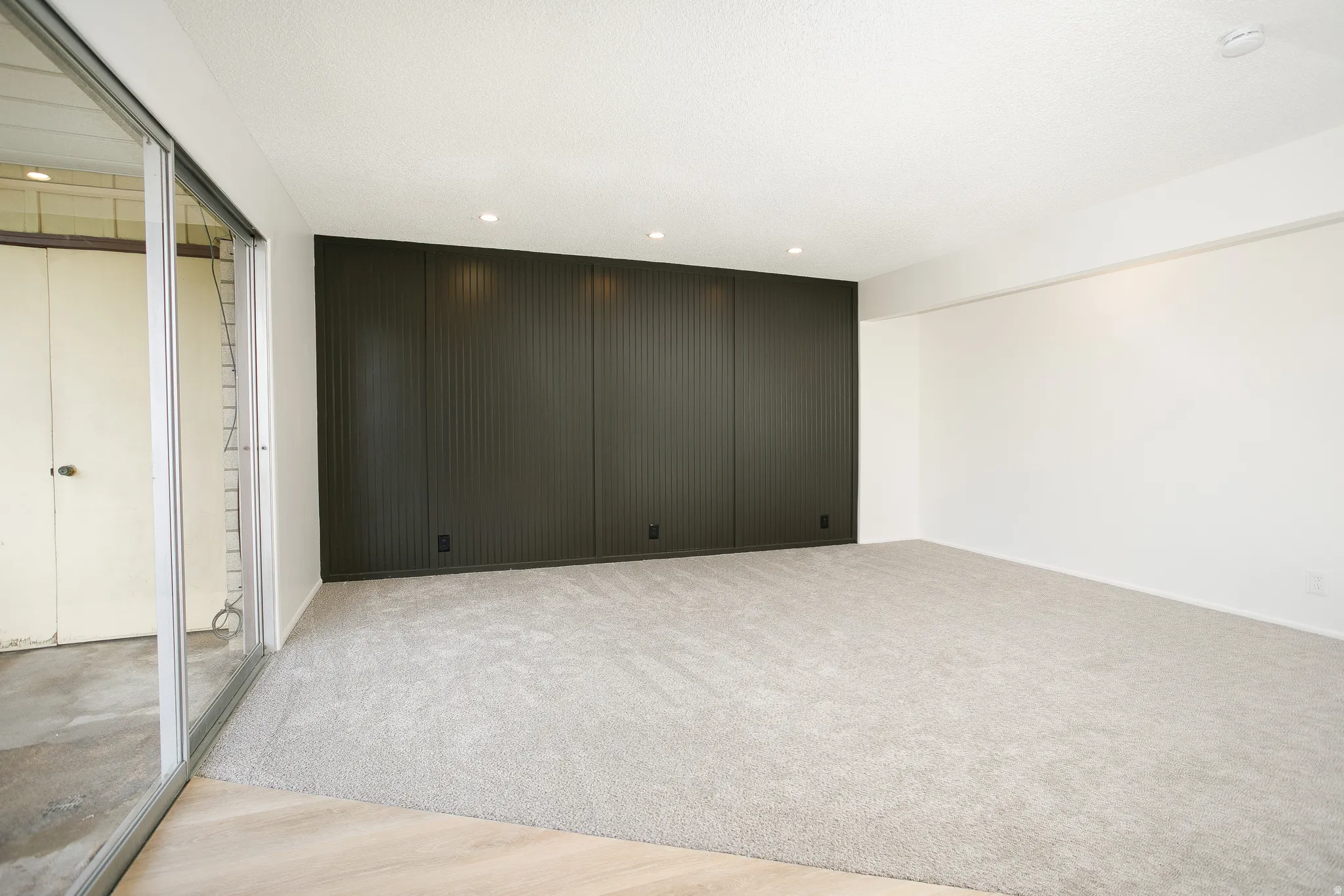 Wall-to-wall carpeting, recessed ceiling lighting, a dark accent wall with vertical paneling, and a sliding glass door