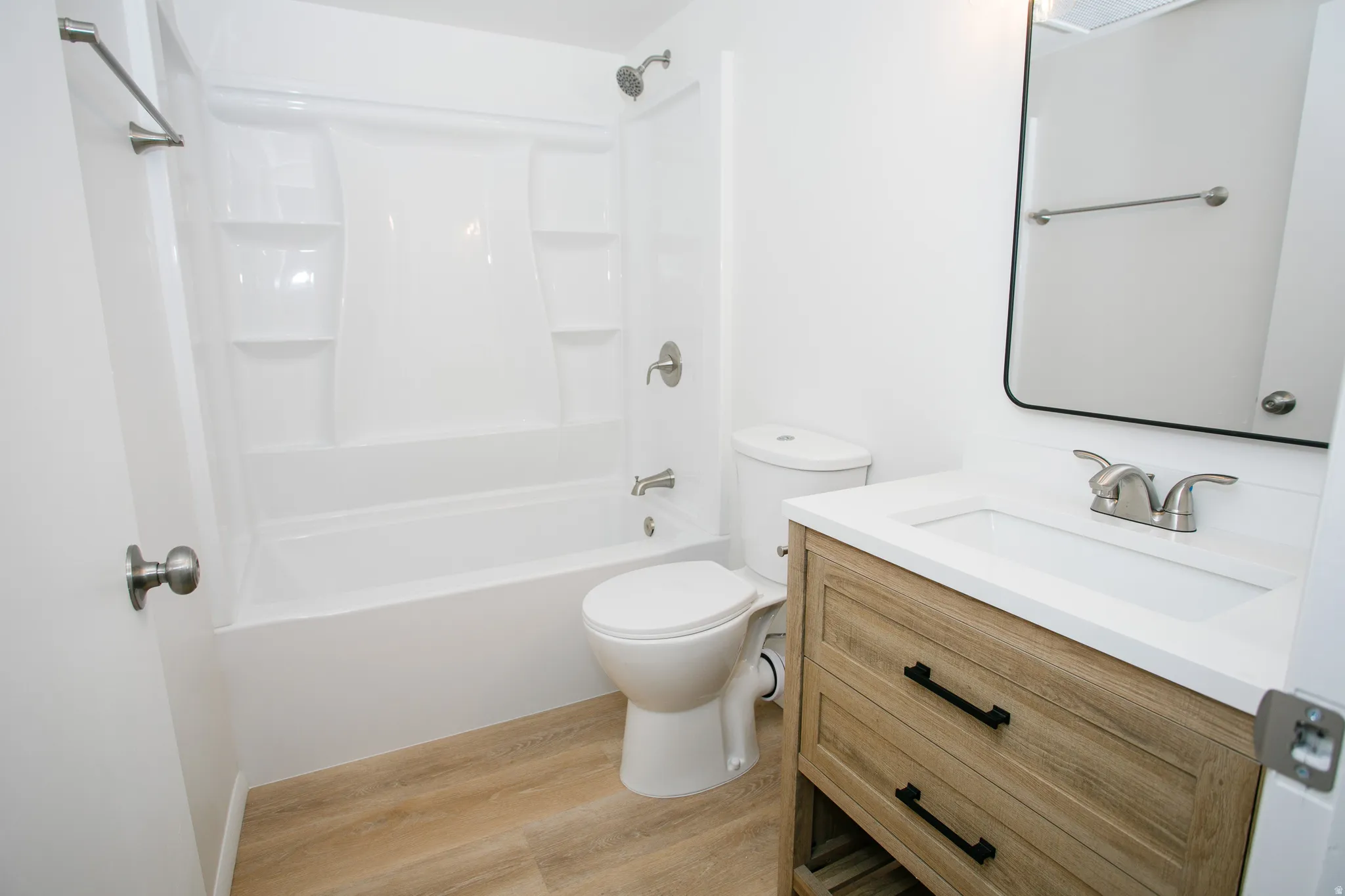 White tub and shower combination with chrome fixtures, wood-finish flooring, and a vanity with a white countertop and black hardware
