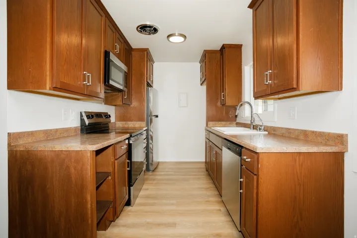 Galley kitchen featuring wood-finish cabinetry, stainless steel appliances, solid surface countertops, and wood-finish flooring