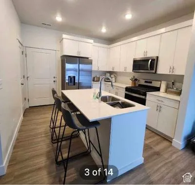 Kitchen featuring a center island with integrated sink, white cabinetry, stainless steel appliances, and wood-finish flooring
