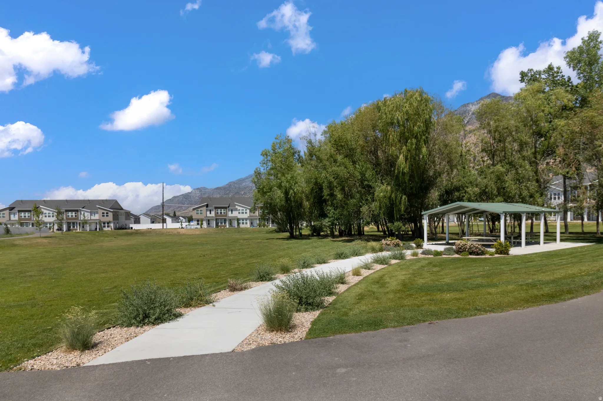 Expansive grassy area with a paved walking path, mature trees, a covered outdoor structure, and distant mountain backdrops