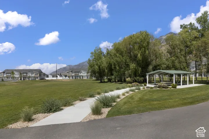 Expansive grassy area with a paved walking path, mature trees, a covered outdoor structure, and distant mountain backdrops