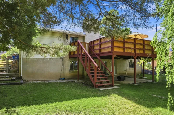 Expansive green lawn with a multi-level deck, exterior stairs, concrete patio, and mature trees