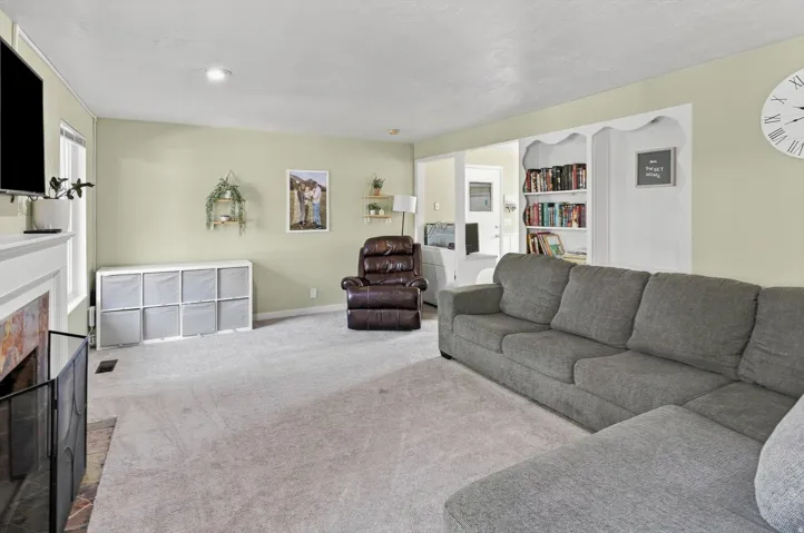 Spacious living area featuring a curved sectional sofa, fireplace with white surround and black insert, light yellow walls, recessed ceiling lighting, built-in shelving with storage cubbies, wall-mounted clock, and carpet flooring throughout the room
