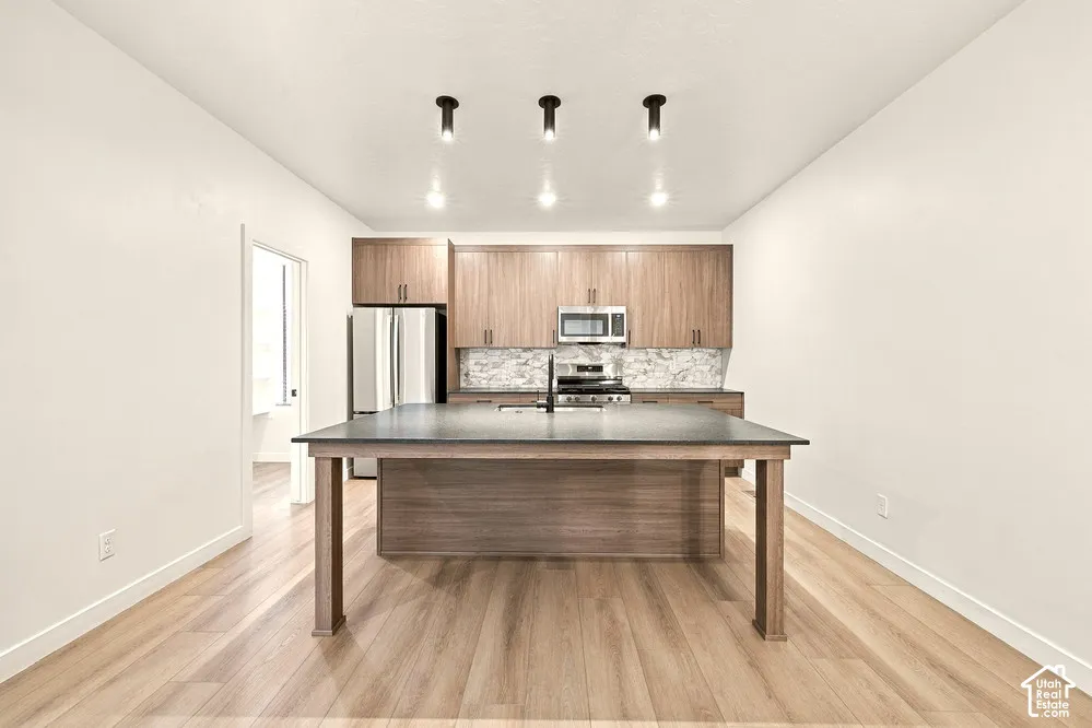 Kitchen featuring wood-finish cabinetry, a large central island with a solid surface countertop, wood-finish flooring, stone-pattern backsplash, and recessed cylinder lighting