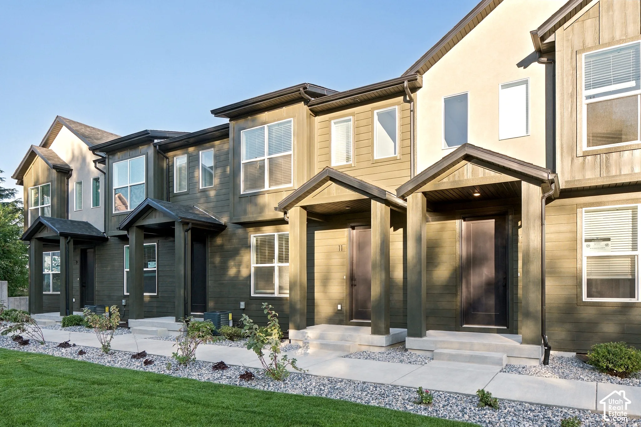 Multi-unit exterior featuring mixed siding, covered entryways with structural support columns, concrete walkways, and manicured landscaping with stone ground cover