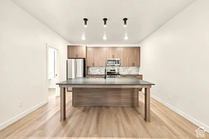 Kitchen featuring wood-finish cabinetry, a large central island with a solid surface countertop, wood-finish flooring, stone-pattern backsplash, and recessed cylinder lighting