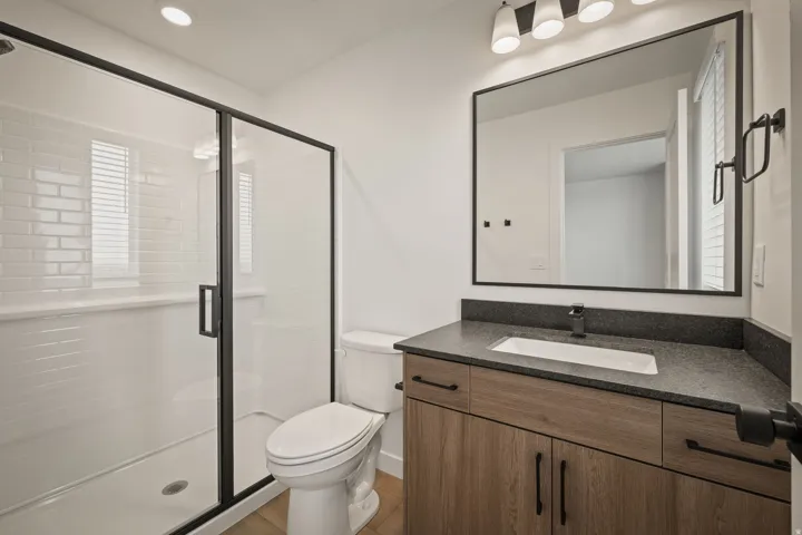 Glass-enclosed shower with subway tile, wood-finish vanity with dark countertop, rectangular undermount sink, and black-framed mirror