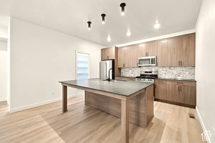 Kitchen featuring wood-finish cabinetry, stone-surface countertops, an island with integrated sink, stone backsplash, and cylindrical ceiling lighting