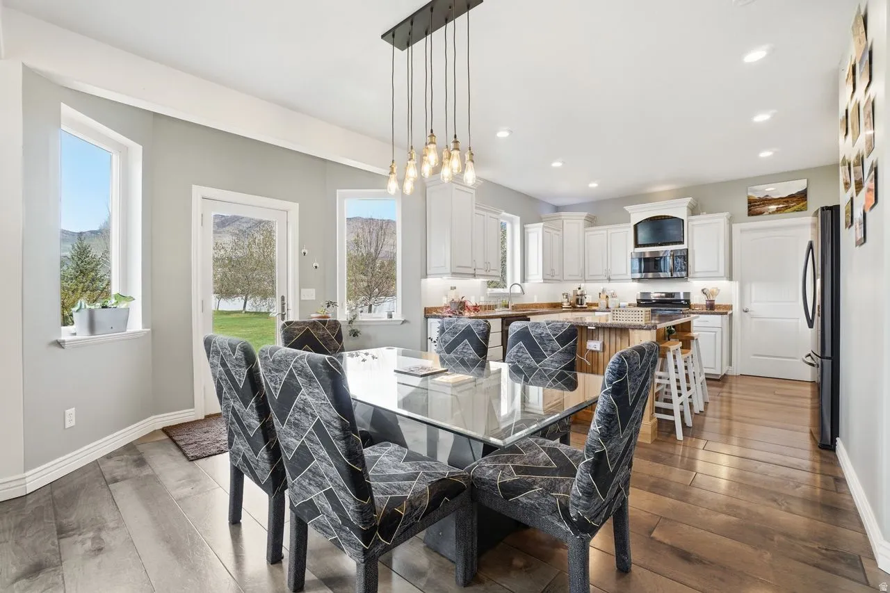 Open-concept dining and kitchen area featuring a glass-top dining table with upholstered chairs, white cabinetry with stainless steel appliances, quartz countertops, a multi-pendant light fixture suspended from the ceiling, hardwood flooring, and expansiv