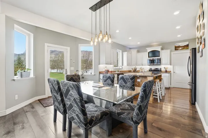 Open-concept dining and kitchen area featuring a glass-top dining table with upholstered chairs, white cabinetry with stainless steel appliances, quartz countertops, a multi-pendant light fixture suspended from the ceiling, hardwood flooring, and expansiv