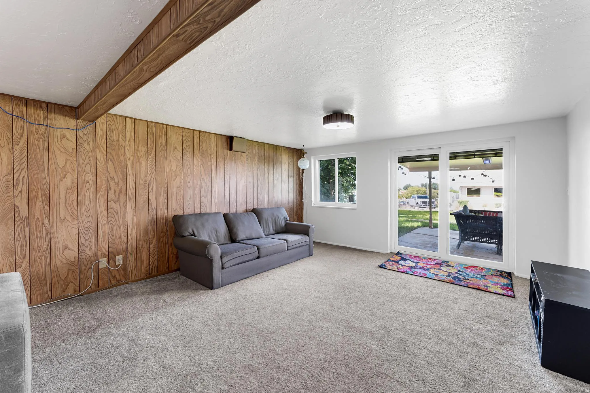 Carpeted flooring, wood-panel accent wall, recessed ceiling beam, dual-pane sliding glass door, and a single window