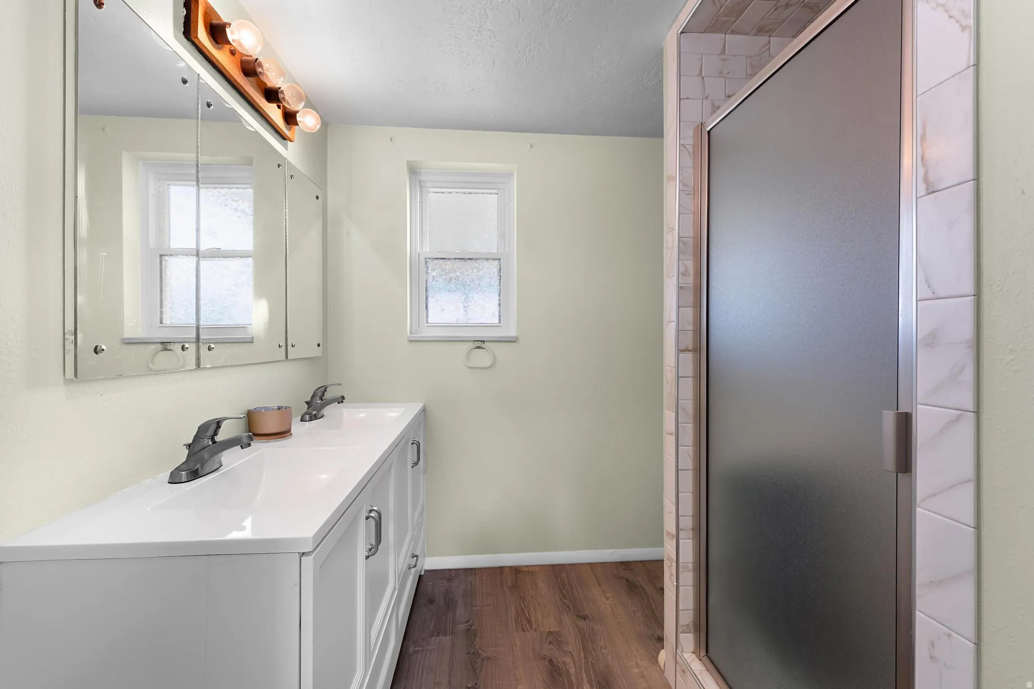 Dual-sink vanity with white countertop, wood-finish flooring, and a shower with a frosted glass door