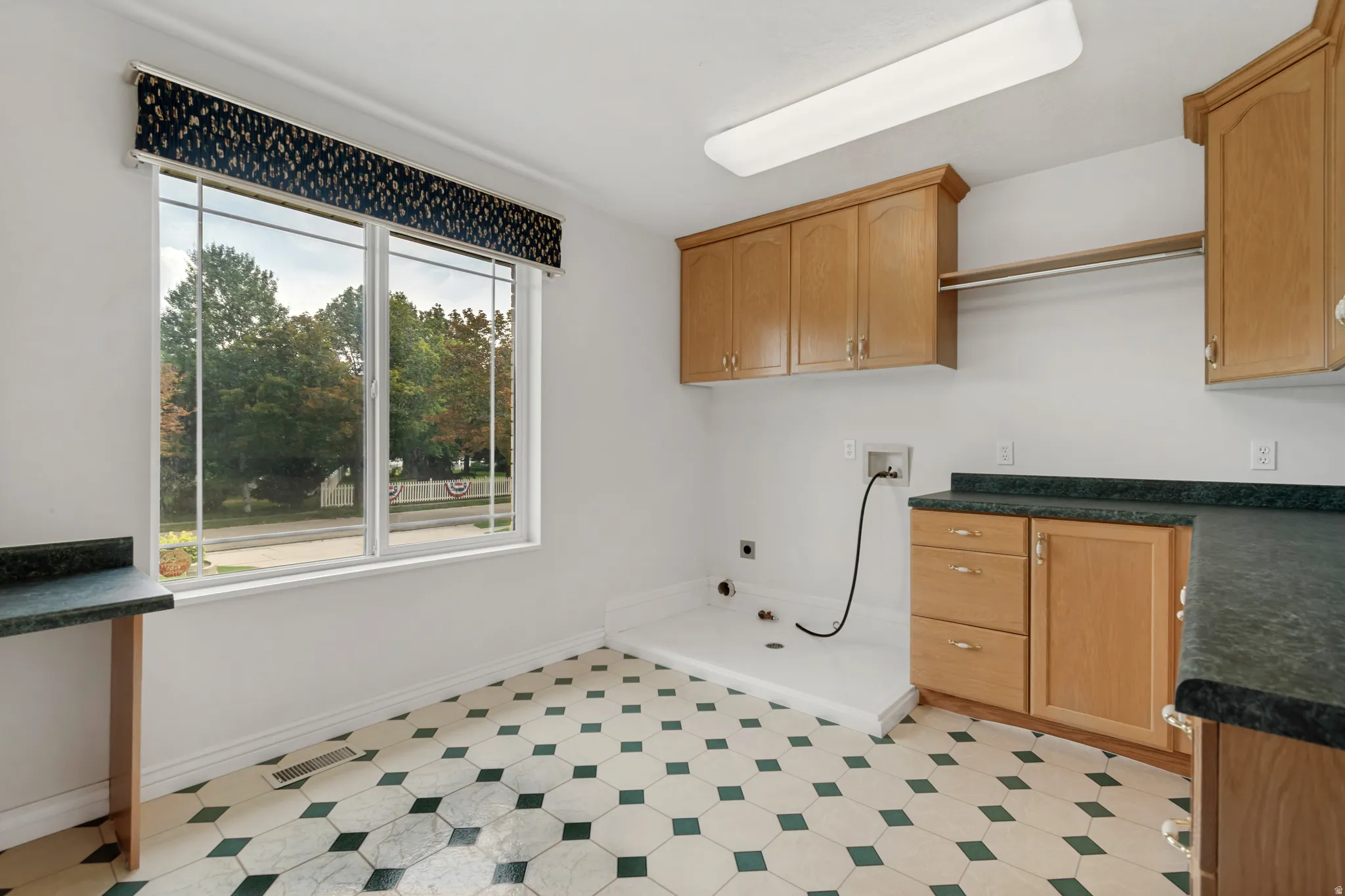 Laundry room featuring a large window, hexagonal tile flooring, light wood cabinetry, dark countertops, and an integrated clothing rod