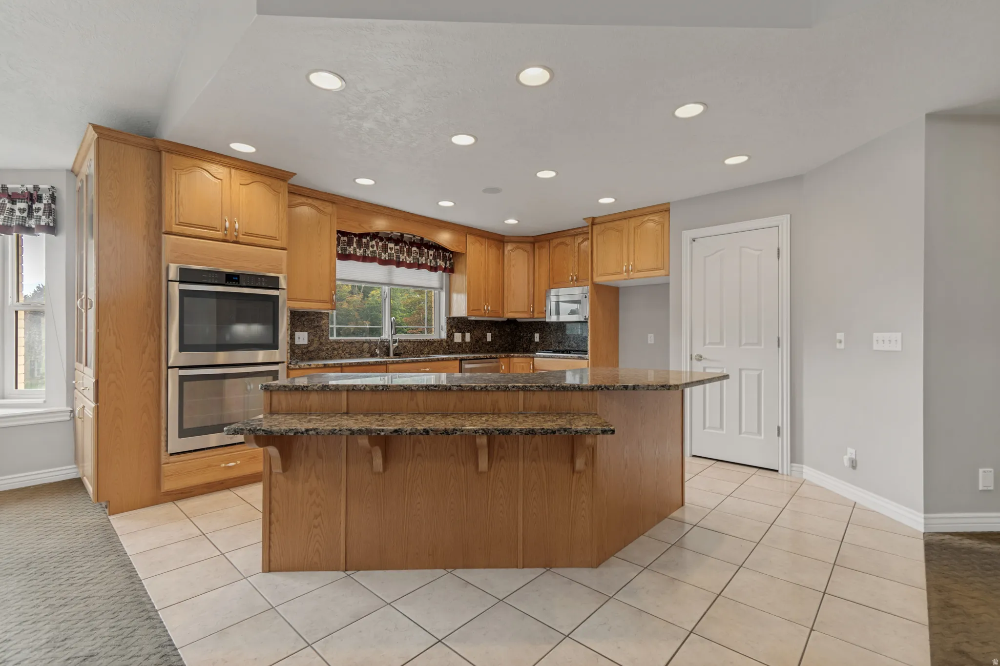Kitchen featuring wood cabinetry, stainless steel double ovens, an island with an extended counter, tiled flooring, and recessed lighting