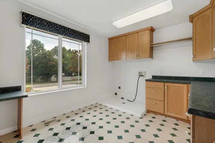 Laundry room featuring a large window, hexagonal tile flooring, light wood cabinetry, dark countertops, and an integrated clothing rod