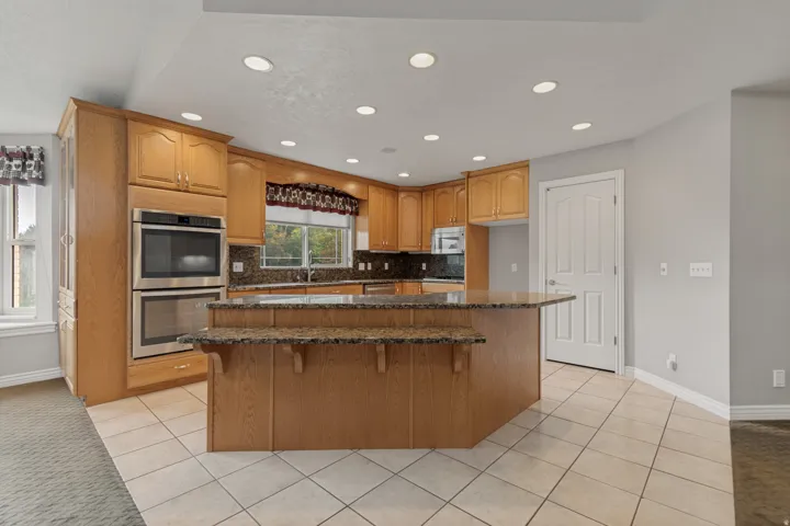 Kitchen featuring wood cabinetry, stainless steel double ovens, an island with an extended counter, tiled flooring, and recessed lighting