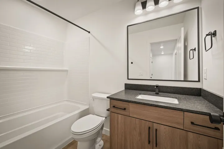 Bathtub with white subway tile surround, wood-finish vanity with dark countertop, integrated rectangular sink, matte black faucet, and rectangular mirror with black frame