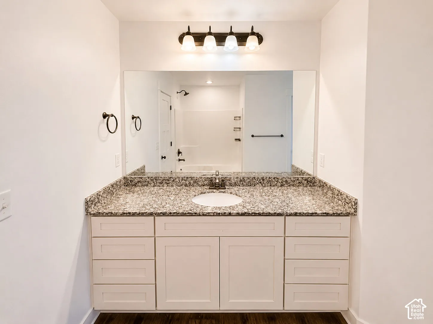 White vanity with integrated sink, granite countertop, wall-mounted mirror, and dark wood-finish flooring