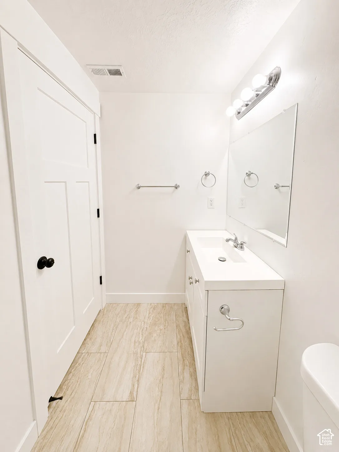 White vanity with integrated sink, rectangular mirror, chrome light fixture, and wood-look tile flooring