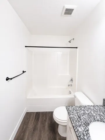 White bathtub/shower combination, dark wood-finish flooring, white vanity with granite-style countertop, and a white toilet