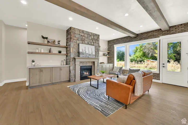 Spacious living area featuring a stone-clad fireplace, wood-finish flooring, exposed ceiling beams, built-in shelving with lower cabinetry, and large windows providing abundant natural light