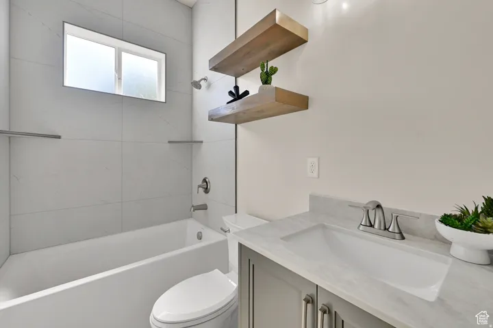 Bathroom featuring a white tub, large format tiled shower surround, frosted window, gray vanity with undermount sink, and light-toned countertop