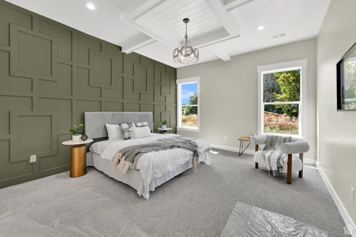 Accent wall with geometric paneling, coffered ceiling with central chandelier, two tall windows with white trim, and light gray wall paint
