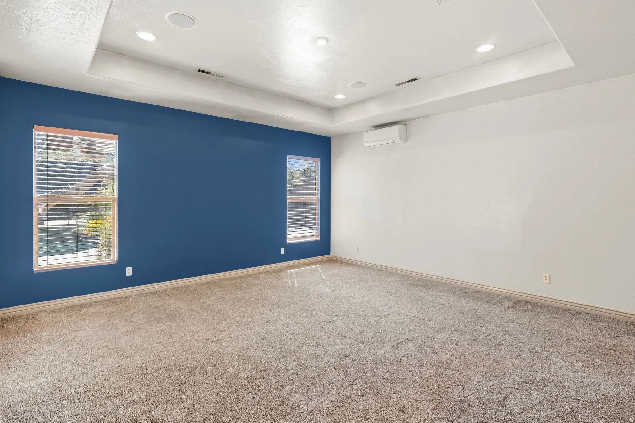Recessed lighting, tray ceiling, two windows with blinds, wall-mounted mini-split system, and neutral carpet flooring