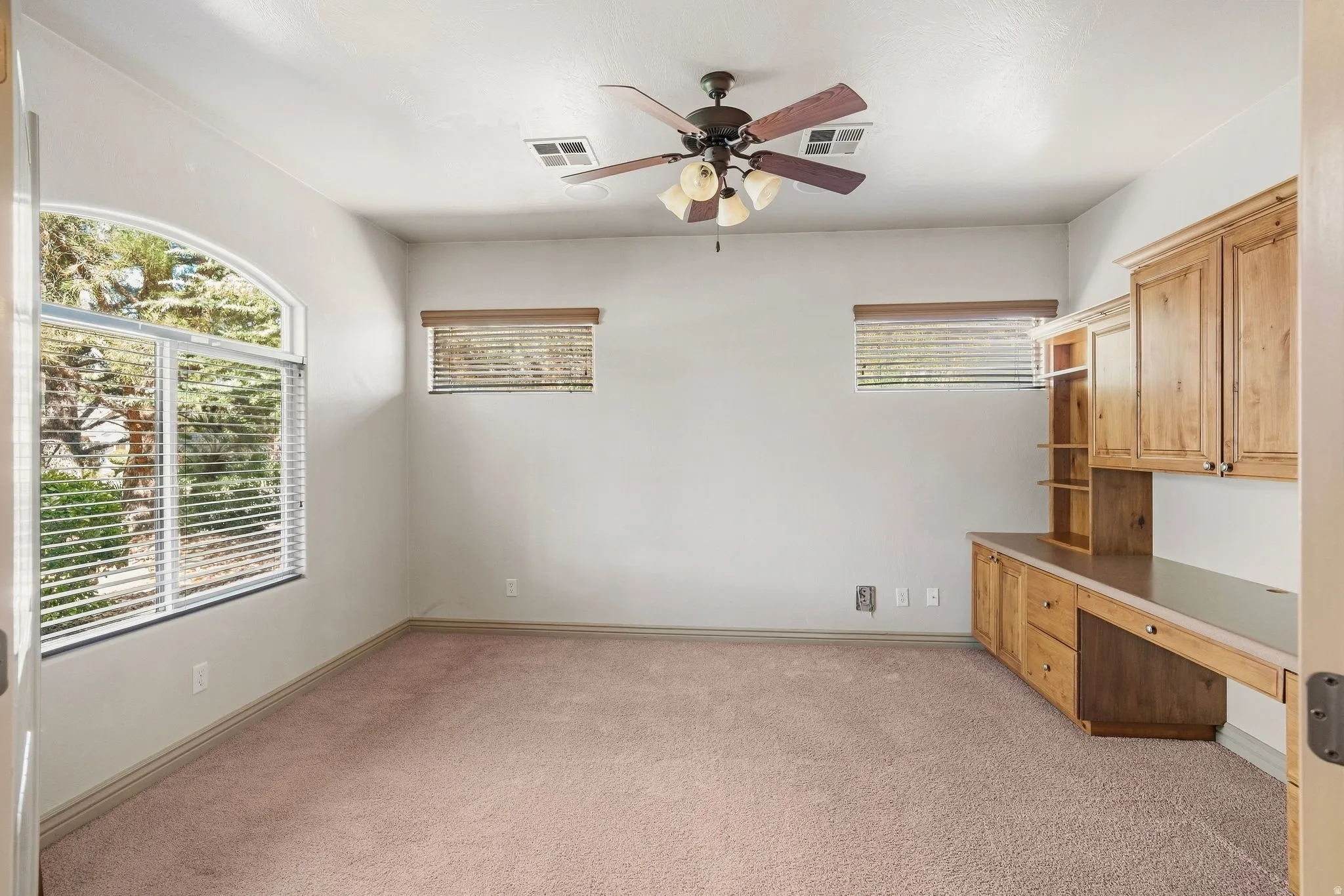 Arched window with blinds, built-in wooden cabinetry, integrated desk space, and a ceiling fan with light fixture