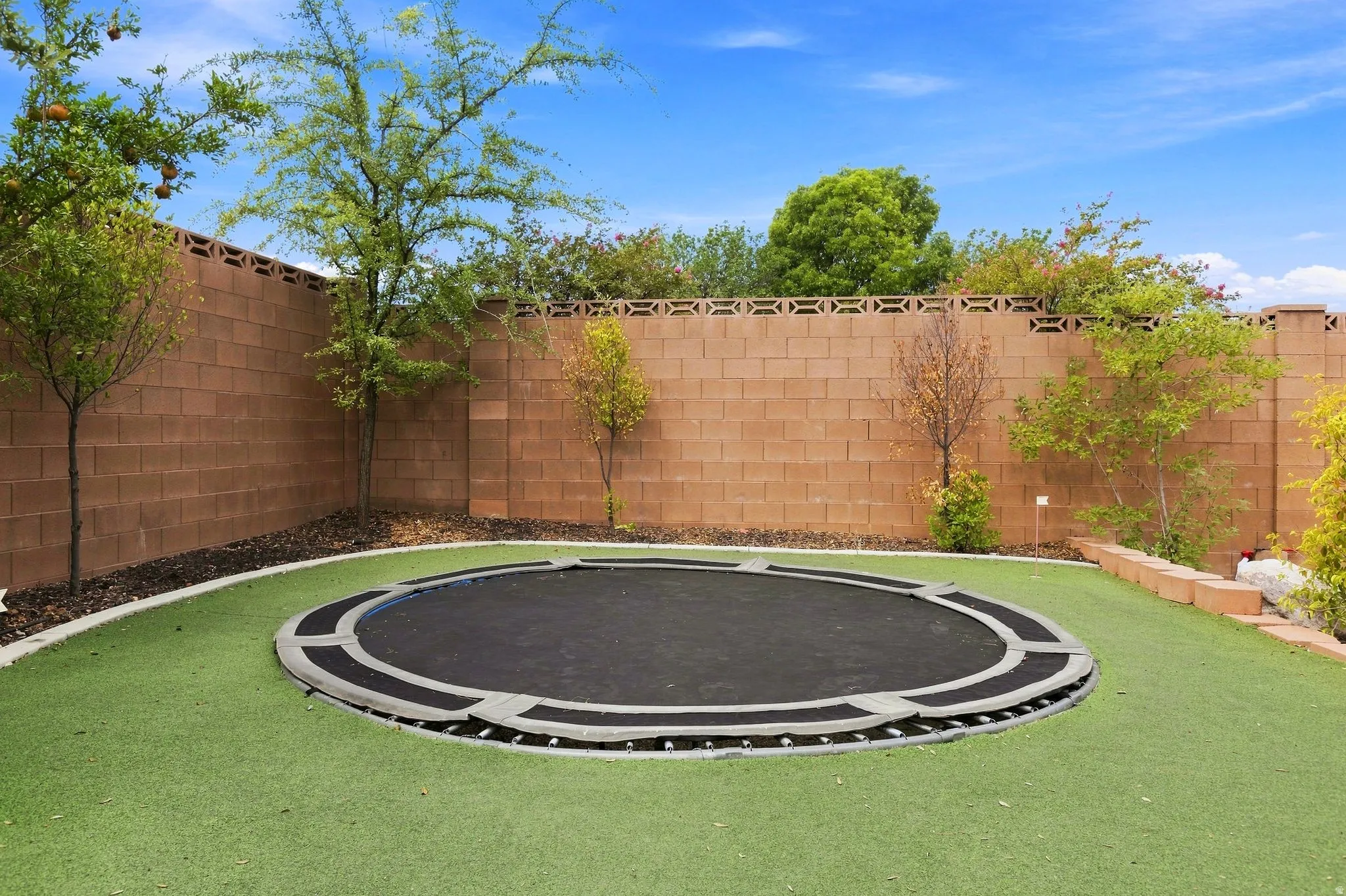 Landscaped backyard featuring artificial turf, perimeter block wall, mature trees, and an in-ground trampoline