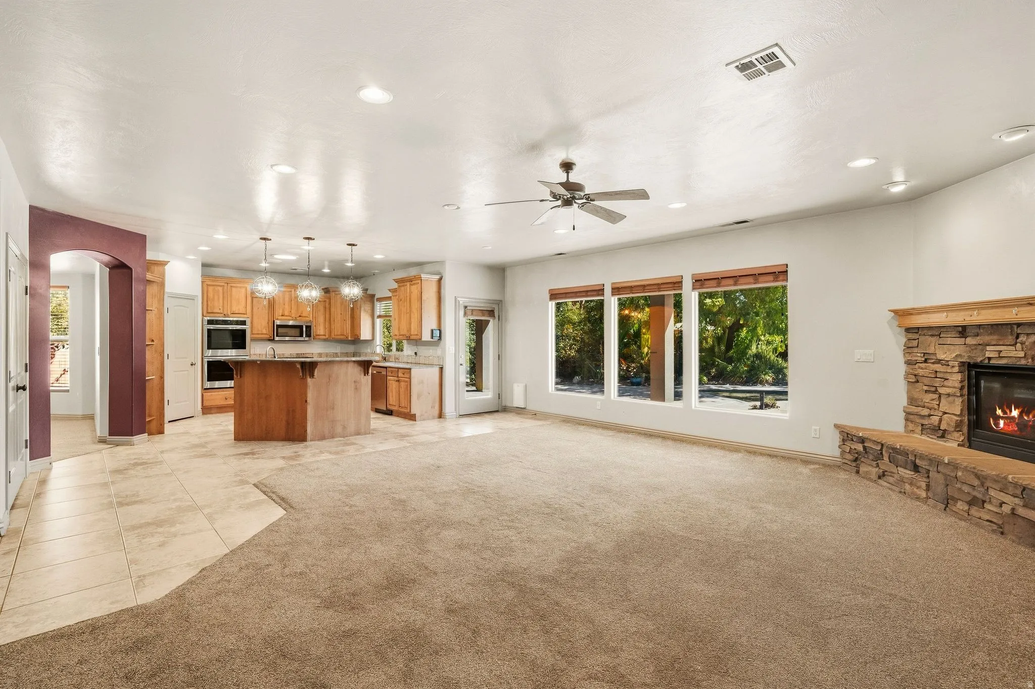 Stone-clad fireplace, recessed lighting, ceiling fan, multiple large windows, and a kitchen with wood cabinetry