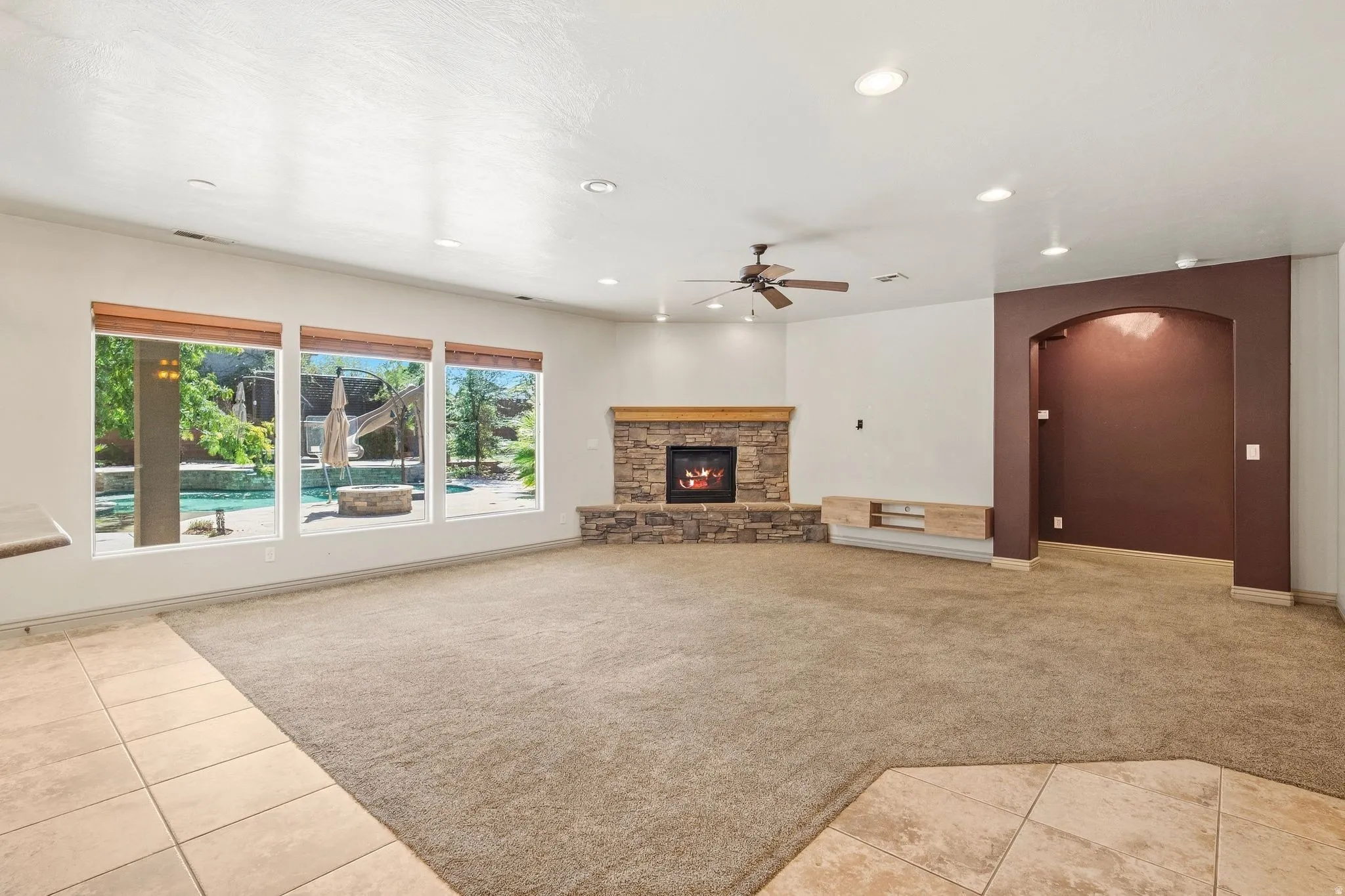 Stone-clad fireplace with a wood mantle, recessed lighting, ceiling fan, and large windows with wood blinds overlooking a backyard with a pool