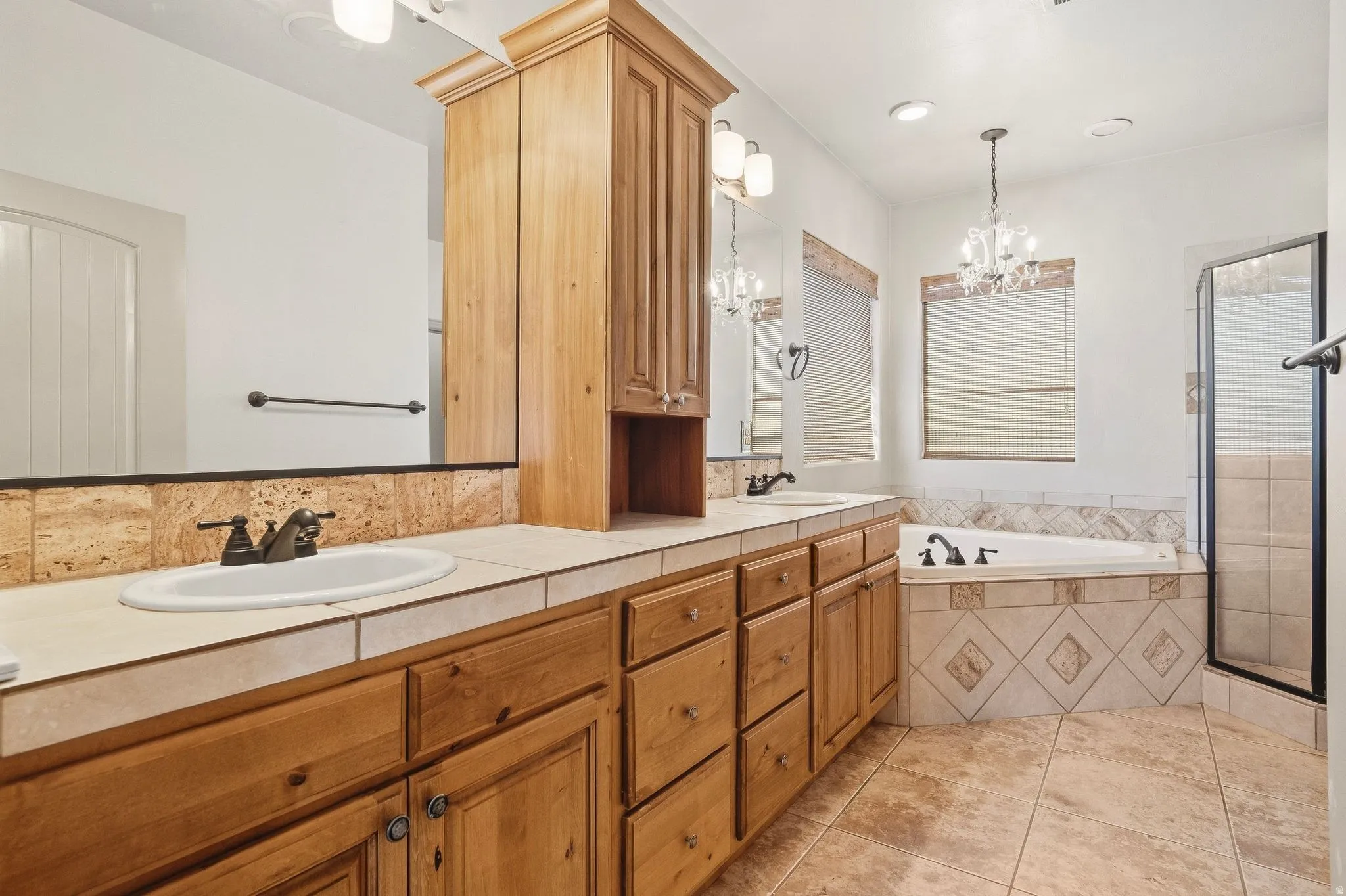 Dual sink vanity with wood cabinetry, tiled countertop, built-in soaking tub, and a separate glass-enclosed shower