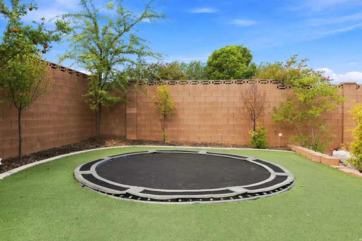 Landscaped backyard featuring artificial turf, perimeter block wall, mature trees, and an in-ground trampoline