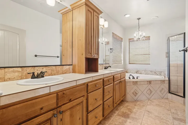 Dual sink vanity with wood cabinetry, tiled countertop, built-in soaking tub, and a separate glass-enclosed shower