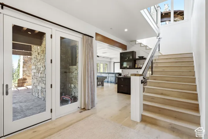 Light wood-finish flooring, white French doors, stone exterior wall, open staircase with metal railing, and white walls