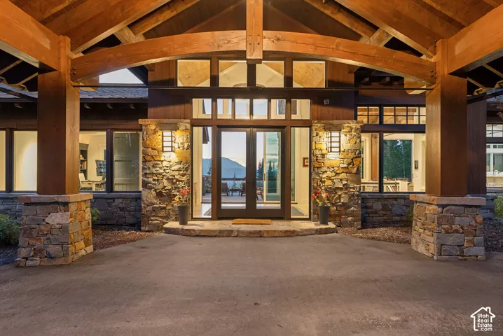 Grand entry featuring exposed timber beams, stone columns, a glass paneled door, exterior wall sconces, and multiple large windows