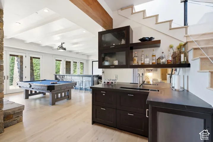 Built-in wet bar with dark cabinetry and countertop, light wood-finish flooring, exposed wood ceiling beams, and a stone-clad architectural element
