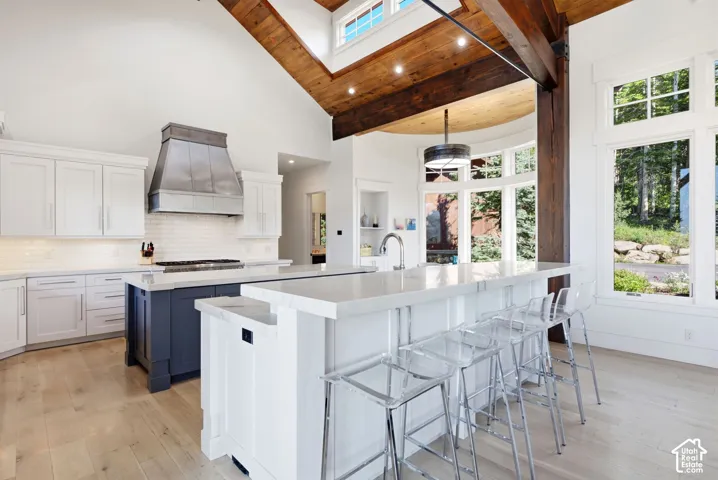 Vaulted ceiling with exposed wood beams, wood-finish flooring, two kitchen islands with solid surface countertops, white cabinetry, and a stainless steel range hood