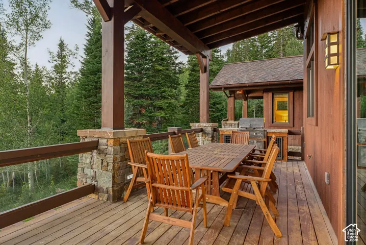 Expansive wood deck with a built-in outdoor kitchen, stone accents, glass panel railing, and wood-paneled overhead coverage