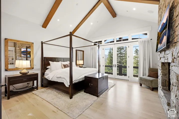 Vaulted ceiling with exposed wood beams, light wood-finish flooring, stone-clad fireplace, and double doors with transom windows