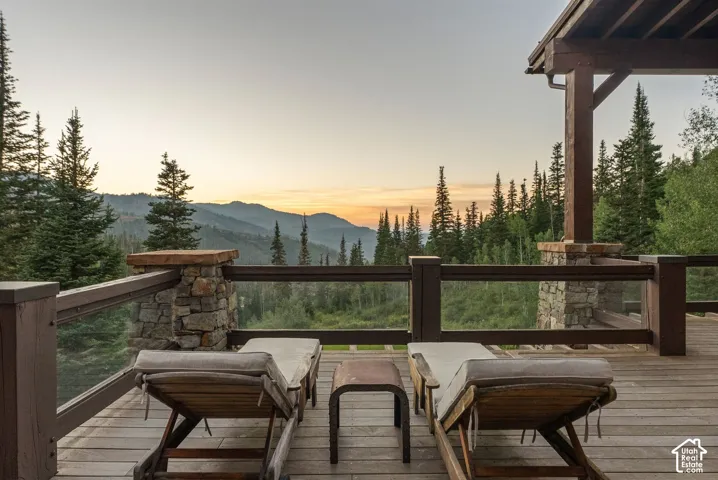 Expansive wood deck with glass panel railing and stone-clad pillars, overlooking a mountain and forest backdrop