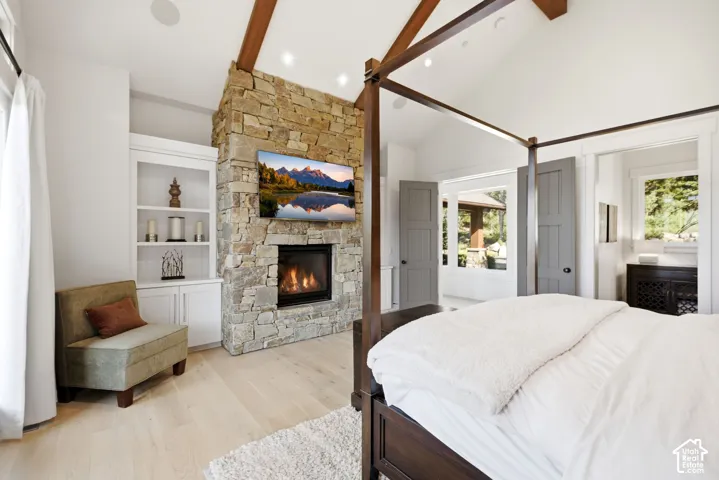 Vaulted ceiling with exposed wood beams, stone-clad fireplace, integrated built-in shelving, light wood-finish flooring, and recessed lighting