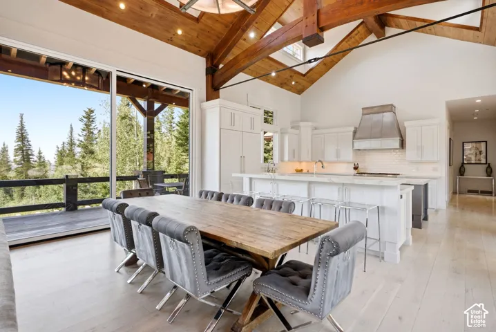 Vaulted ceilings with exposed timber beams, expansive sliding glass doors, contemporary kitchen with white cabinetry and island, stainless steel range hood, and light wood-finish flooring