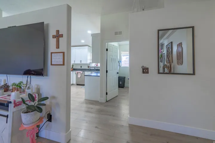 Open floor plan with wood-finish flooring throughout, featuring a bright living area with wall-mounted flat-screen television, kitchen with white cabinetry and stainless steel appliances visible in the adjoining space, and framed wall art