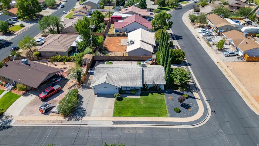Aerial view showcasing a single-story residence with a light-colored roof, expansive manicured lawn with mature trees, curved driveway with dark asphalt, and established landscaping throughout the corner lot