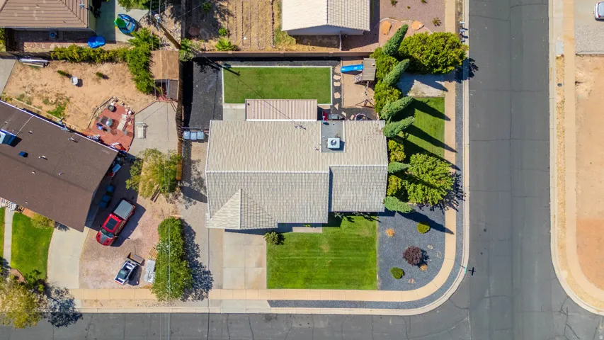 Aerial view showcasing a residence with a light-colored shingled roof, manicured lawn, mature landscaping with established trees, concrete driveway, and defined hardscaped perimeter