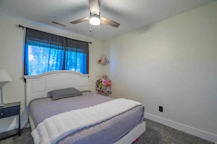 Comfortable secondary bedroom featuring a curved headboard bed frame, soft purple bedding, carpeted flooring, and large window with blue curtains allowing natural light