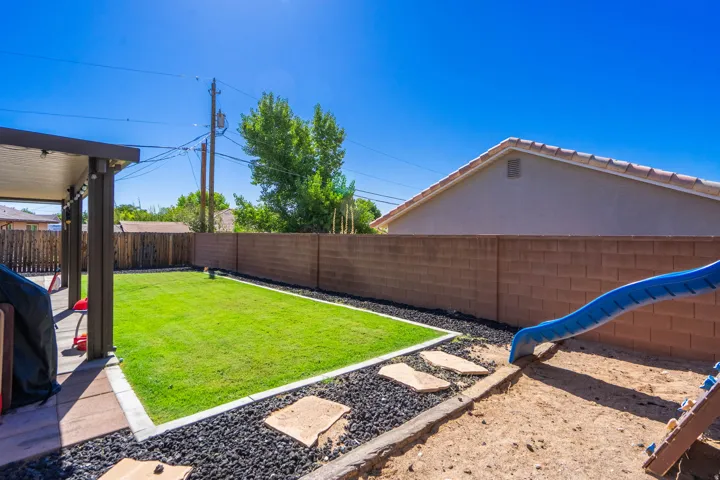 Covered patio with integrated lighting, manicured lawn bordered by black decorative rock, and concrete block privacy wall